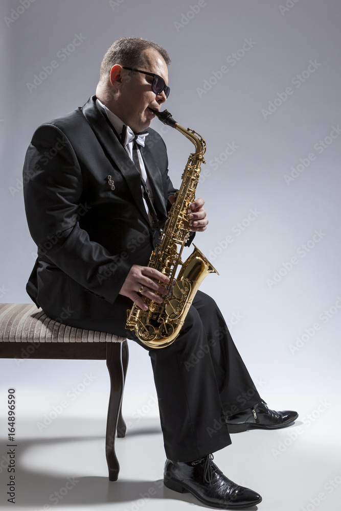 Fototapeta premium Music Concepts. Portrait of Mature Relaxed and Thoughful Caucasian Saxophone Player in Sunglasses Playing the Saxophone While Sitting on Chair in Studio Environment.