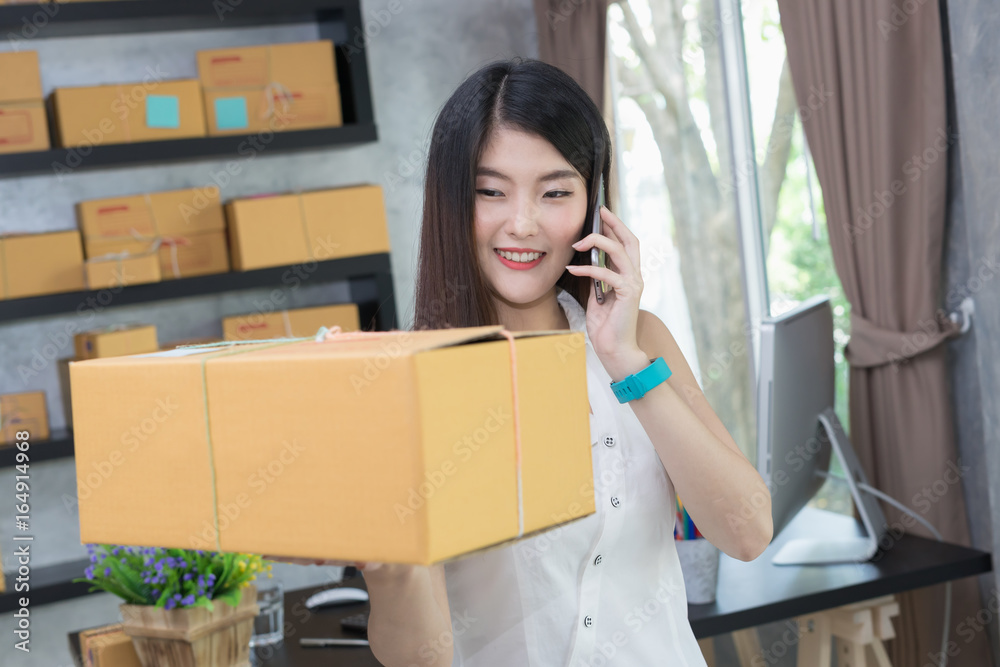 Asian woman entrepreneur is using phone and showing the box packing at home workplace Asian woman entrepreneur is using phone and showing the box packing at home workplace