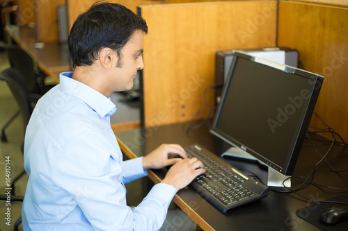 Closeup portrait, young handsome man in blue shirt typing away, browsing digital computer, isolated background of indoors library cubicle background