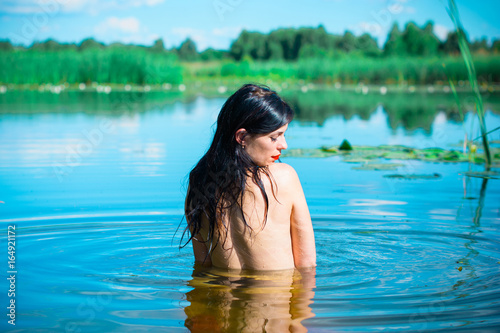 Mermaid girl with dark hair on a blue lake. Sexy and thin figure, red lids and pale skin. Inspire woman bathed in water and seduces