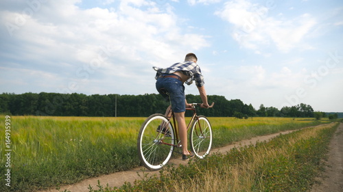 Fototapeta Naklejka Na Ścianę i Meble -  Young man riding vintage bicycle at the rural road over field. Sporty guy cycling along country trail outdoor. Male cyclist riding bike in the countryside. Healthy active lifestyle Slow motion