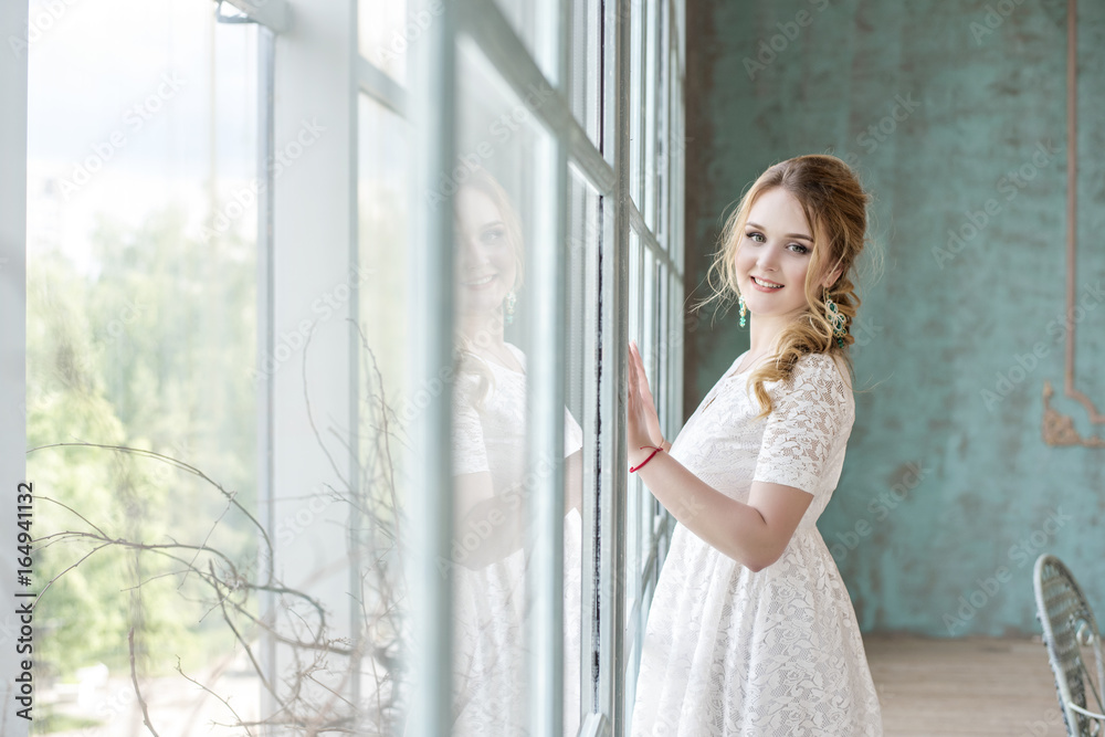 Young very beautiful girl is standing in a white dress near a large window in a bright room Young very beautiful girl is standing in a white dress near a large window in a bright room