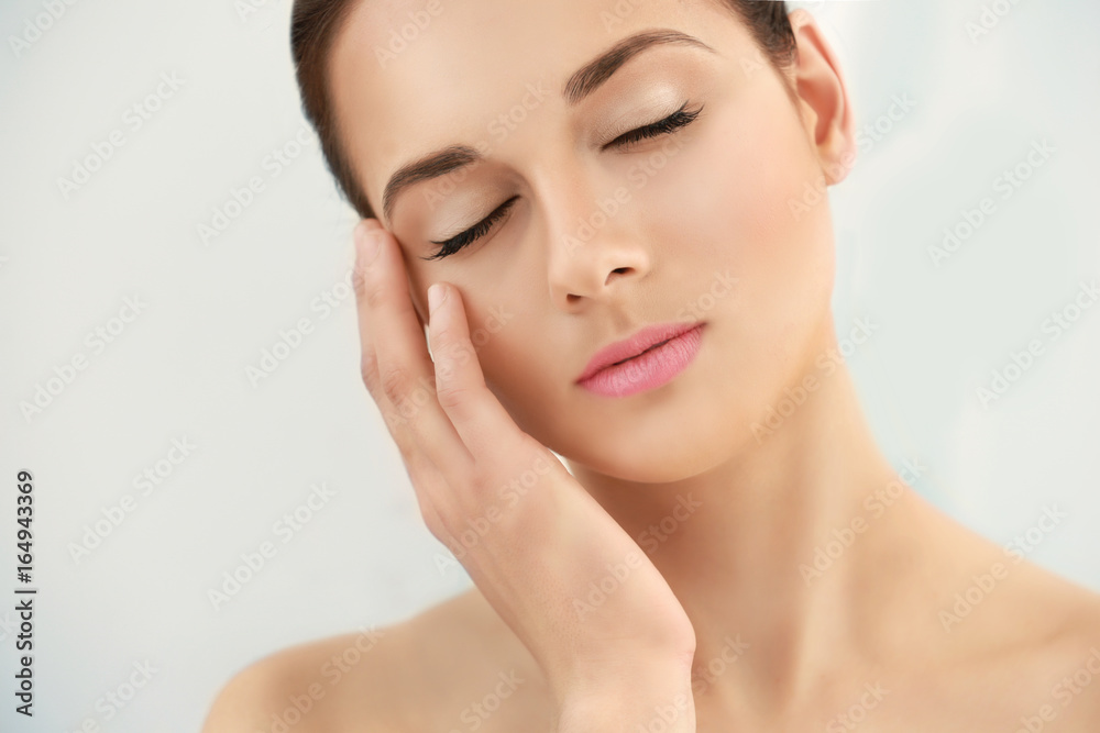 Closeup view of beautiful young woman with natural lips makeup touching face on light background Closeup view of beautiful young woman with natural lips makeup touching face on light background