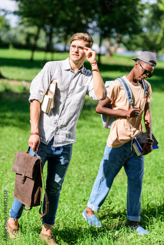 stylish young multiethnic men holding books and backpack while walking together in park