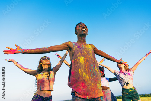 happy young multiethnic friends standing with open arms at holi festival of colors