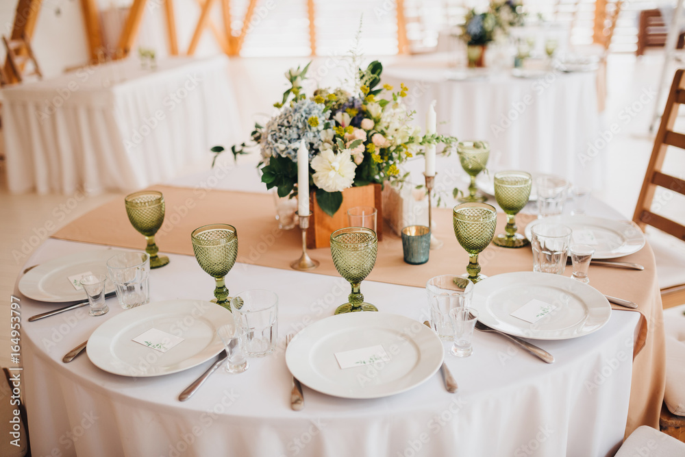 In the wedding banquet area there are wooden tables with tablecloths, on tables there are compositions of flowers and greens, candles, cutlery, on plates lie napkins with name cards