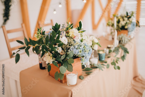 In the wedding banquet area there are wooden tables with tablecloths  on tables there are compositions of flowers and greens  candles  cutlery  on plates lie napkins with name cards