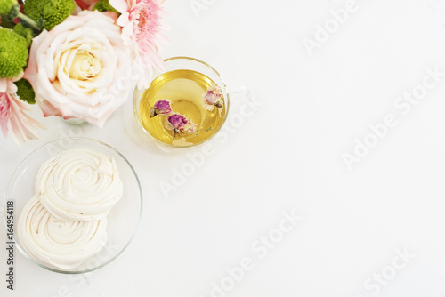 A cup of healthy herbal tea with dried roses. Beautiful fresh flowers, French meringue cookies on light marble table, top view. Pink roses and gerberas on a female work desk. 