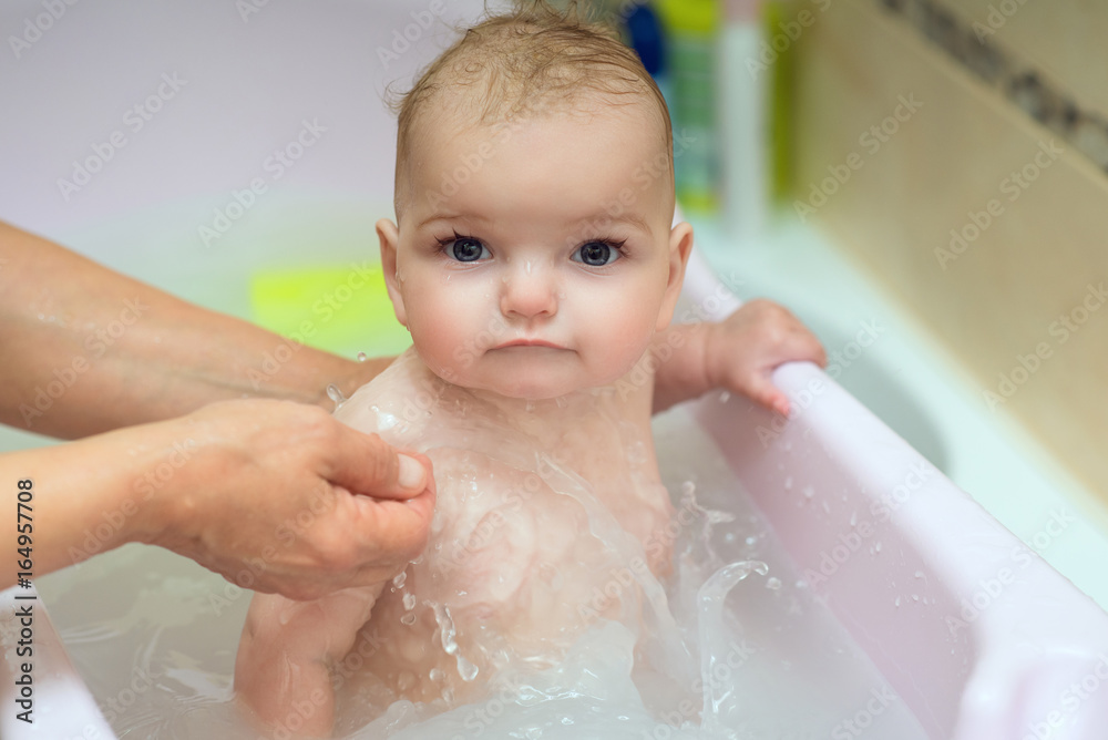 Close up portrait of a beautiful baby girl taking emollient bath, her mom’s hands pouring water on her. Indoor shot Close up portrait of a beautiful baby girl taking emollient bath, her mom’s hands pouring water on her. Indoor shot