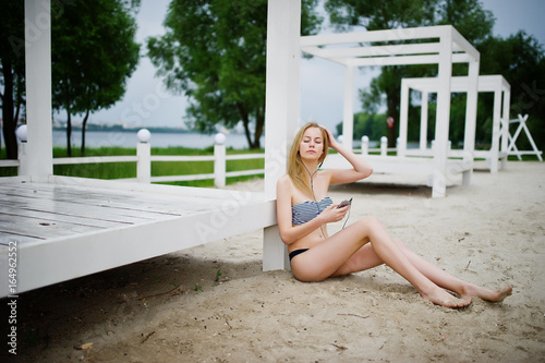 Portrait of a gorgeous young girl in bikini sitting next to the gasebo on sand and posing. photo