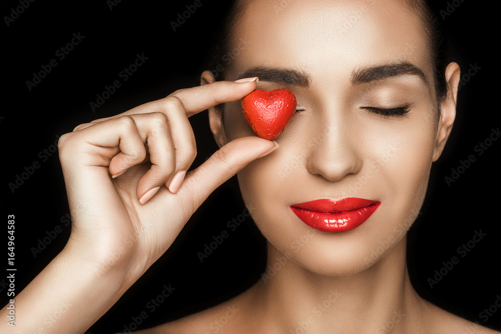 attractive woman with closed eyes holding red heart shaped candy, isolated on black attractive woman with closed eyes holding red heart shaped candy, isolated on black