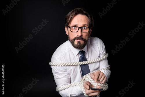 Canvastavla scared middle aged businessman tied with rope using smartphone and looking at ca