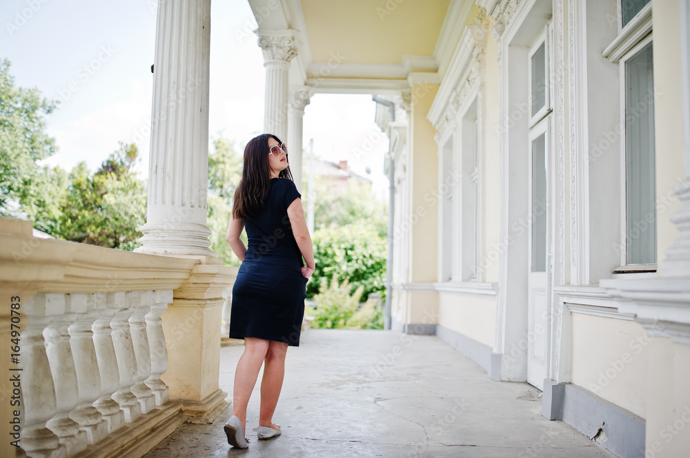 Fototapeta premium Brunette girl at black dress, sunglasses posed against old vintage house, at street of city.