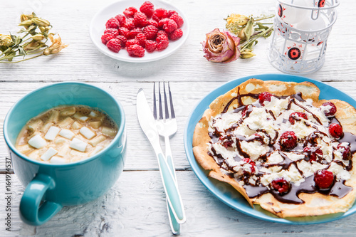 Homemade pancakes or Russian pancakes with chocolate sauce, whipped cream and raspberries on a plate on a white wooden background.