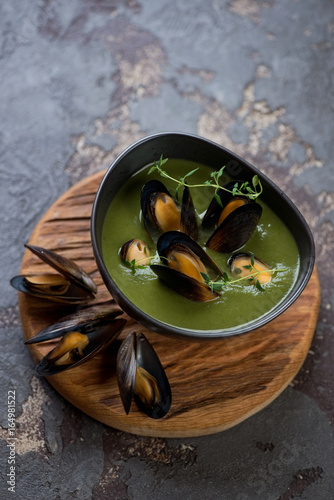 Wooden serving board with a bowl of spinach soup with mussels, brown stone surface, studio shot