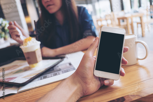 Mockup image of a man's hand holding white mobile phone with blank black screen in modern cafe and blur woman in background