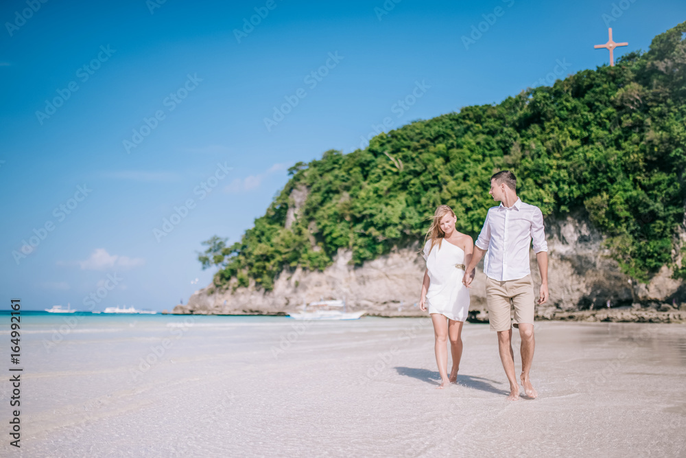 Beautiful couple having fun and walking along a white sandy beach with blue sky and turquois water on the background. Men and women are happy, they looking to each other and smiling together.