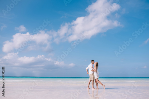 Young man is kissing a beautiful long hair girl in white dress on the white sand beach with a tropical sea and blue sky with clouds on the background. Summer vacation concept.
