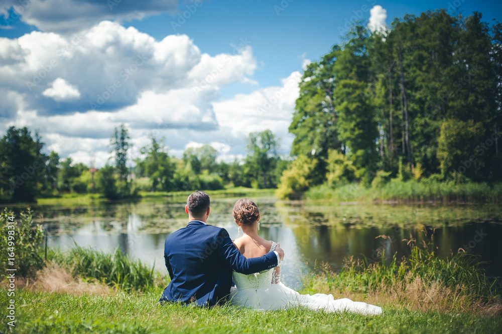 Married couple sitting by the lake. Bride and groom hugging on the grass during sunny day. Married couple sitting by the lake. Bride and groom hugging on the grass during sunny day.