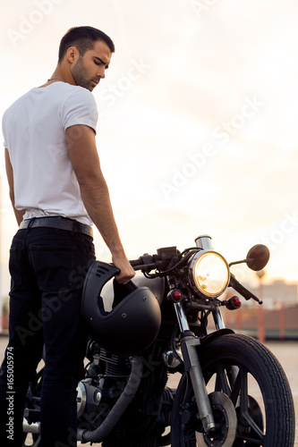 Tela Sporty biker handsome rider male in white blank t-shirt walk to classic style cafe racer motorbike on rooftop at sunset