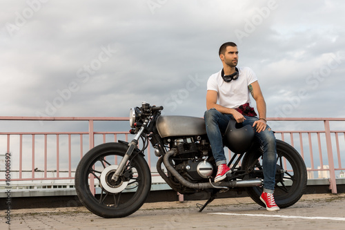 Rider man with beard and mustache in red sneakers and white t-shirt sit on classic style biker cafe racer motorcycle. Bike custom made in vintage garage. Brutal fun urban lifestyle. Outdoor portrait.