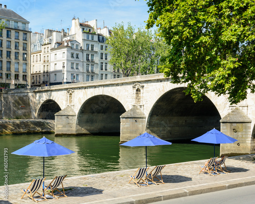 Three blue parasols with blue and white striped deck chairs in the sun on the bank of the river Seine with typical parisian bridge and buildings in the background. photo