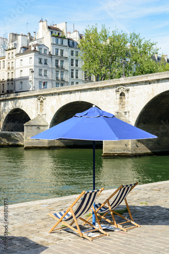 Two blue and white striped deck chairs in the sun under a blue parasol on the bank of the river Seine with typical parisian bridge and buildings in the background. photo