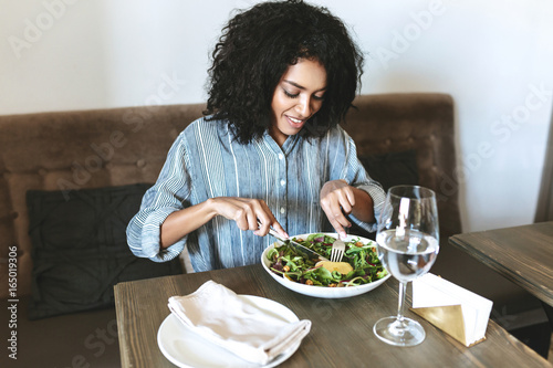 Young African American girl eating salad in restaurant. Beautiful girl with dark curly hair sitting at cafe and eating salad. Portrait of smiling lady that have lunch in restaurant