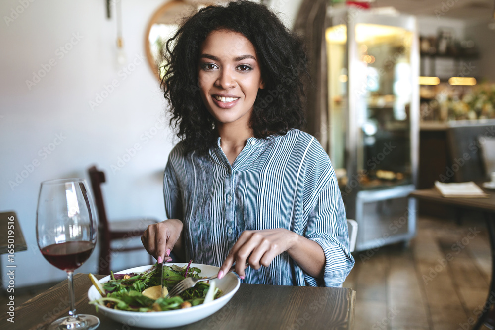 Young African American girl with dark curly hair sitting in restaurant. Smiling girl looking in camera with glass of red wine and salad on table at cafe. Portrait of lady eating salad in restaurant