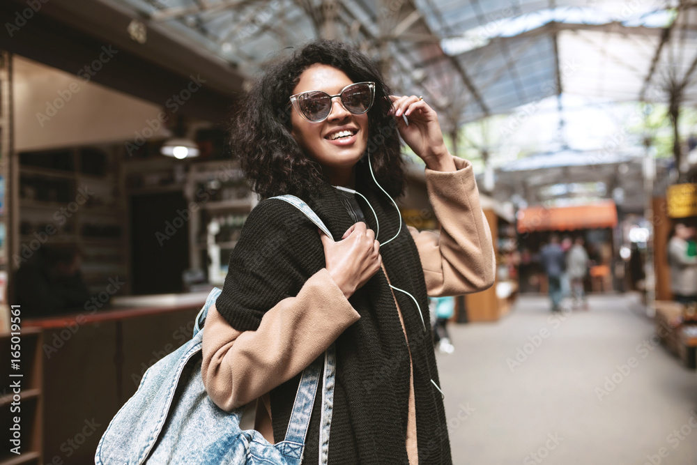 Cool African American girl wearing sunglasses and earphones. Smiling girl  with dark curly hair looking in camera on street. Nice girl in coat,scarf  and bag on her shoulder walking around street Photos