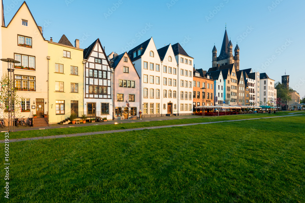 Houses and park in Cologne, Germany. Many of them are colourful, they are facing a public park with green grass and some trees. There is a Cologne bell tower on back.