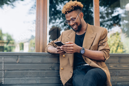 A portrait of African American man in jacket and glasses with smartphone sitting near the windows