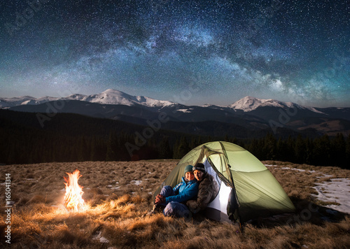 Young couple tourists having a rest in the camping at night, sitting near campfire, looking to the camera under beautiful starry sky and milky way. On the background snow-covered mountains