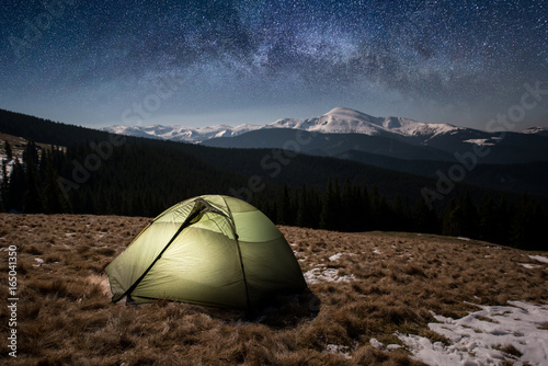 Fototapeta Naklejka Na Ścianę i Meble -  Night camping. Illuminated tourist tent under beautiful night sky full of stars and milky way. On the background snow-covered mountains and forests