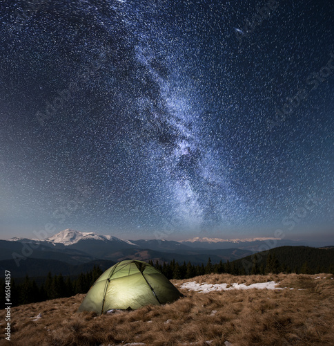 Night camping. Illuminated tourist tent under beautiful night sky full of stars and milky way. On the background snow-covered mountains and forests