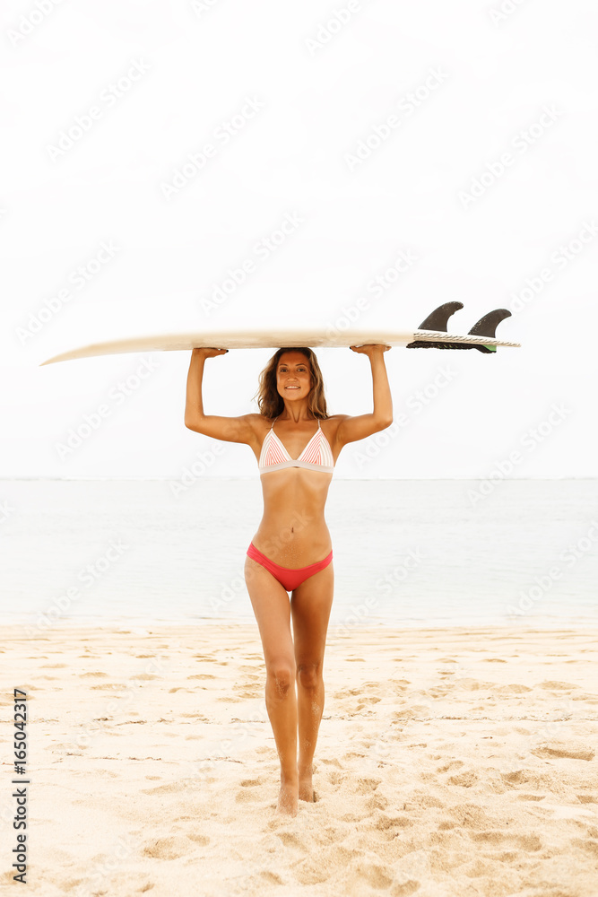 Beautiful fit surfing girl in sexy bikini swimwear with white surf  shortboard surfboard blank board at sunrise or sunset on a beach smile to  camera. Vacation concept. Summer holidays. Tourism, sport. Stock