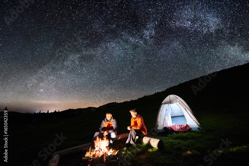 Night camping in the mountains. Man and woman tourists have a rest at a campfire near illuminated tent under incredible night sky full of stars and milky way. Low light