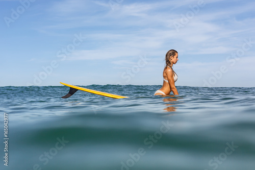 Beautiful surfing girl in sexy bikini sit on a front of big longboard surf board and we see one single fin of her surfboard on sunrise or sunset. Modern active sport lifestyle and summer vacation.