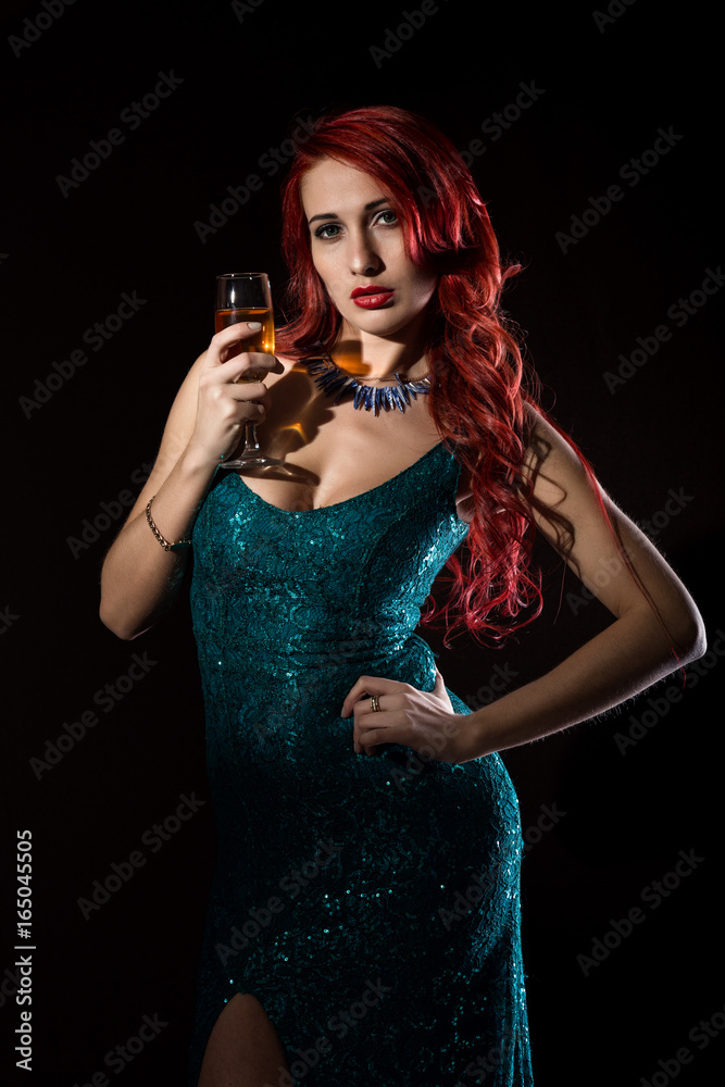 Naklejka premium Young redhead sexy woman in blue elegant dress holding wineglass with champagne and poses in a dark.
