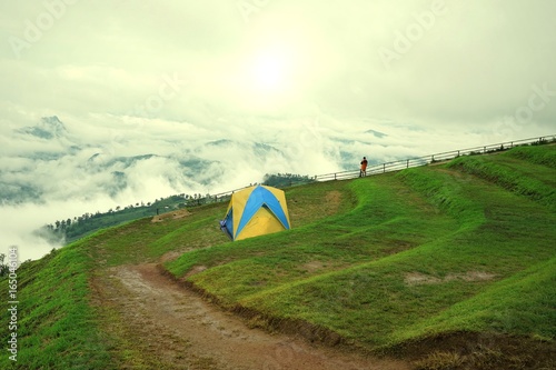 Man in orange jacket looking at view of mountain landscape with waves of fog and cloudy sky. Blue yellow camping tents on mountain peaks at Phu Tub Berg, Phetchabun province, Thailand