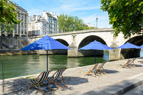Canvas Print Three blue parasols with blue and white striped deck chairs in the sun on the bank of the river Seine with typical parisian bridge and buildings in the background