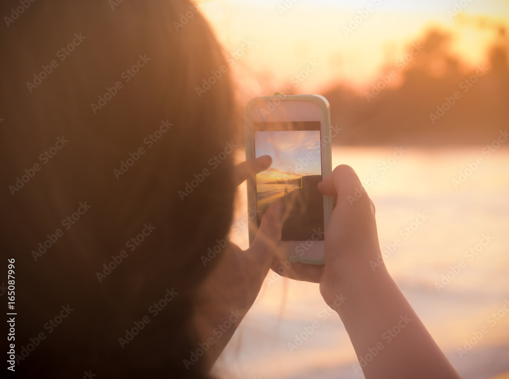 Fototapeta premium Young Woman Takes Photo At Beach on Her Smart Phone at Sunset