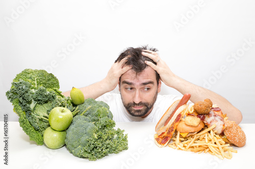 Diet decision and nutrition choices dilemma between healthy good fresh fruit and vegetables or greasy cholesterol rich fast food with a puzzled man trying to decide what to eat. photo