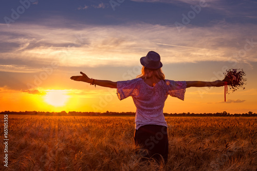 Background girl in a hat with a bouquet of wildflowers in one hand looks at the evening contras sunset standing on the field of ripe wheat and another hand showing peace