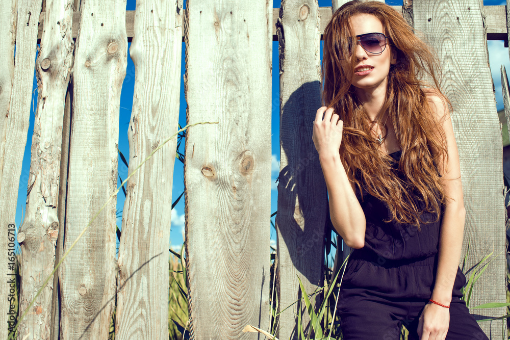 Obraz premium Young beautiful model with long chestnut hair wearing black jumpsuit and trendy aviator sunglasses standing at the old shabby fence made of boards. Copy-space. Outdoor shot