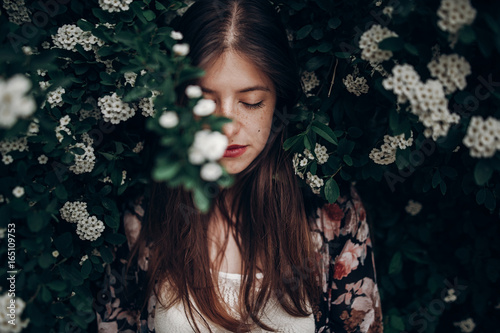 sensual calm portrait of beautiful hipster woman in blooming bush with white flowers of spirea. boho girl sensual portrait in floral modern clothes in greenery. space for text