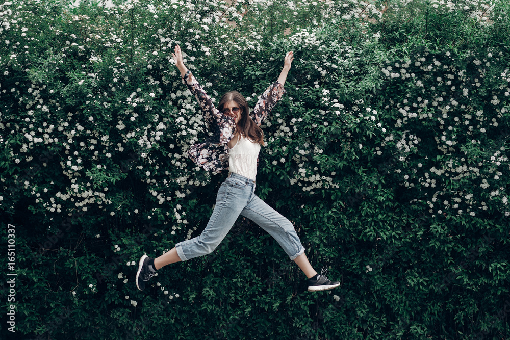 happy hipster woman in sunglasses having fun on background of blooming bush with white flowers of spirea. boho girl jumping and smiling in modern clothes, emotional moment. space for text