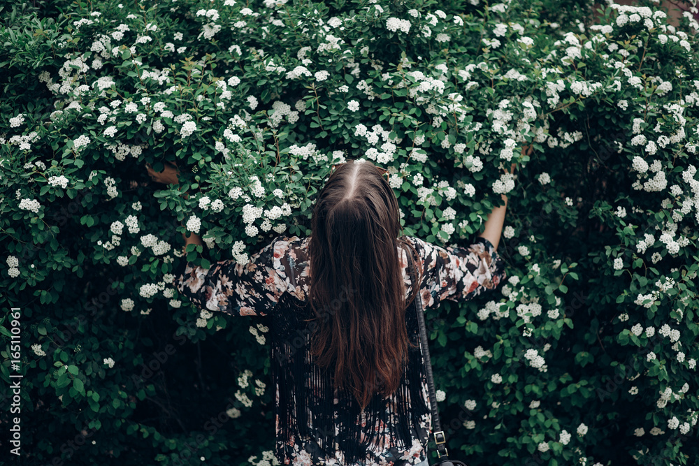 back of happy hipster woman holding blooming bush with white flowers of spirea. boho girl posing in floral clothes. space for text. spring summer time