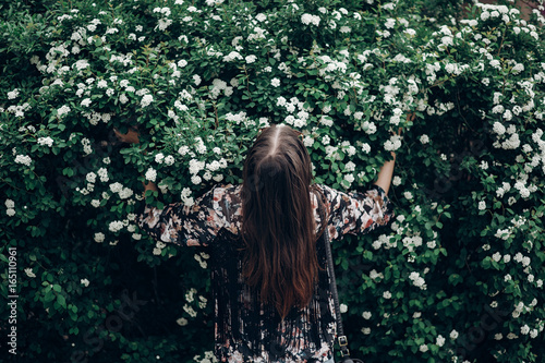 Fototapeta Naklejka Na Ścianę i Meble -  back of happy hipster woman holding blooming bush with white flowers of spirea. boho girl posing in floral clothes. space for text. spring summer time