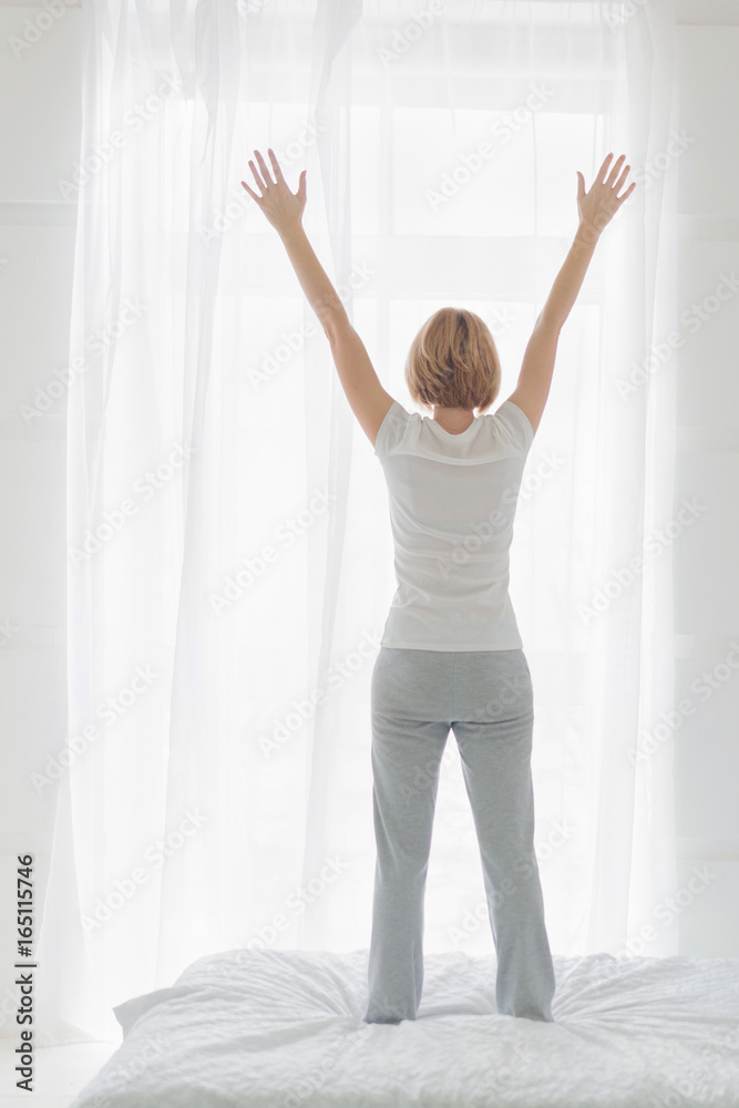Young woman standing on bed in bedroom with hands raised up in the morning. Rear view Young woman standing on bed in bedroom with hands raised up in the morning. Rear view